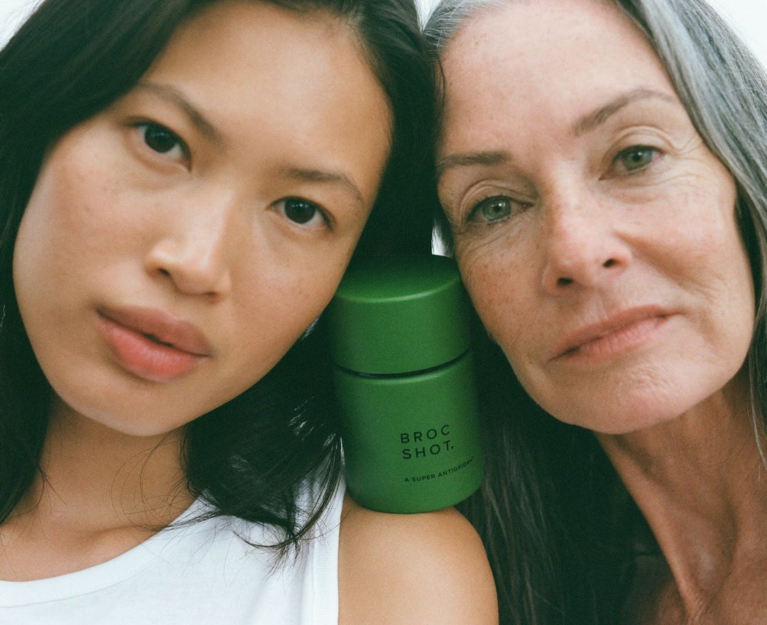 Two women pose with a green supplement jar on the shoulder labeled "BROC SHOT" and "A SUPER ANTIOXIDANT". 