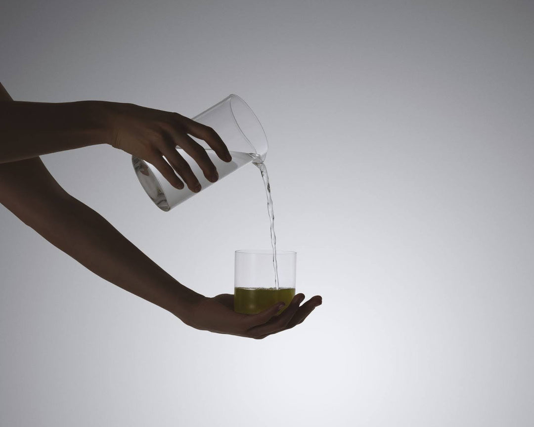 Hands pouring water from a clear pitcher into a glass containing green liquid.