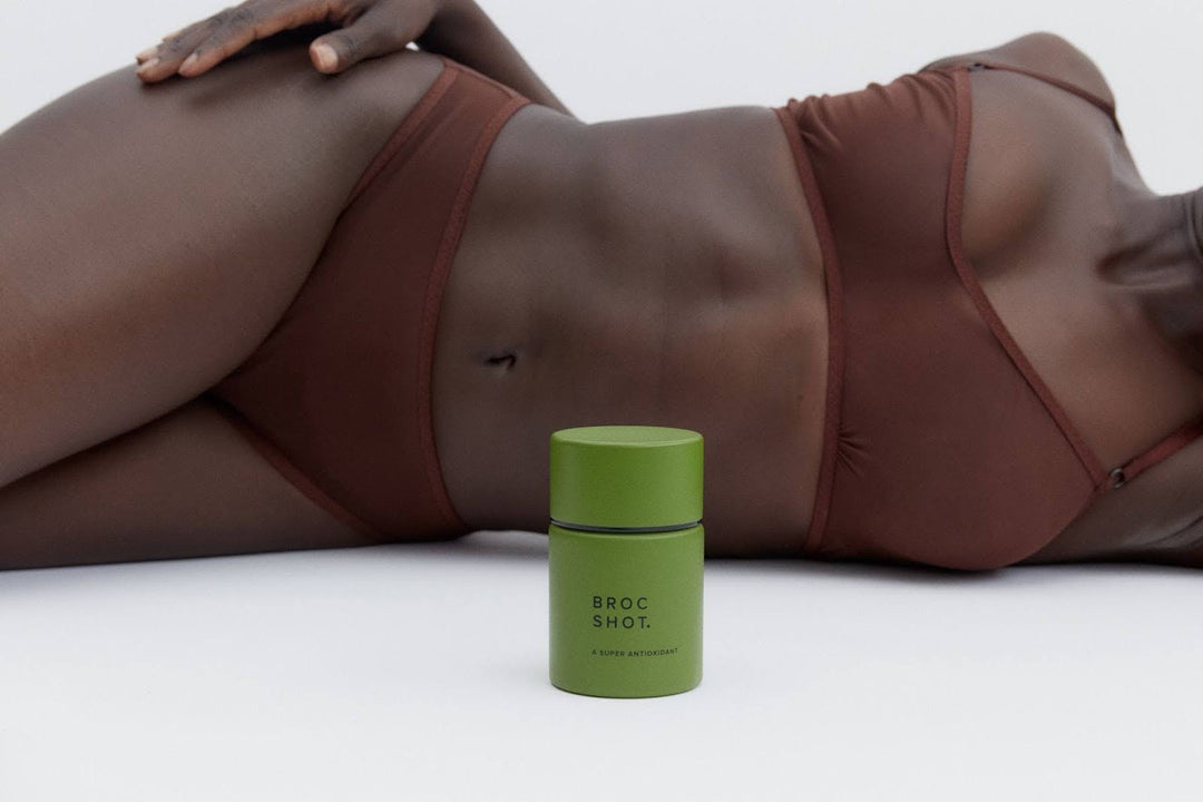 Small green container labeled BROC SHOT. A SUPER ANTIOXIDANT in front of a reclining woman in brown bikini.