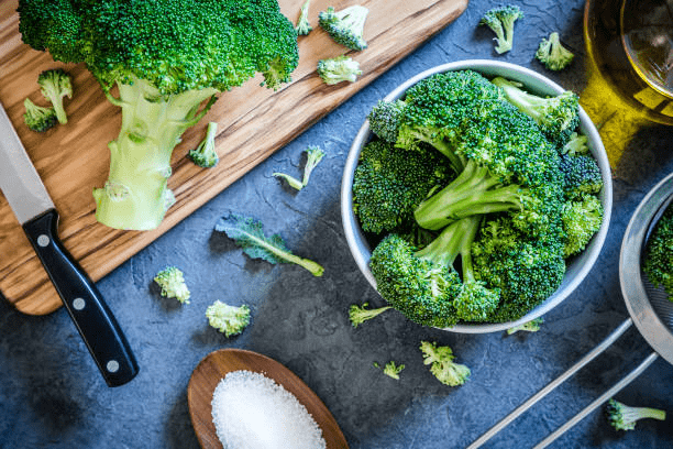 Fresh broccoli florets on a cutting board and in a bowl, with a knife, wooden spoon of salt, and olive oil.