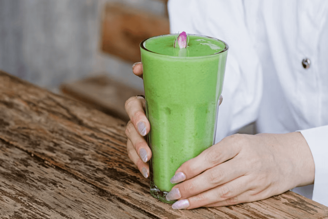 Hands with pale pink nail polish holding tall glass of bright green smoothie topped with a small pink flower petal.