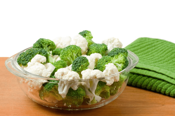 Three-quarter view of a glass bowl filled with raw broccoli and cauliflower florets on a wooden table with a folded green kitchen towel