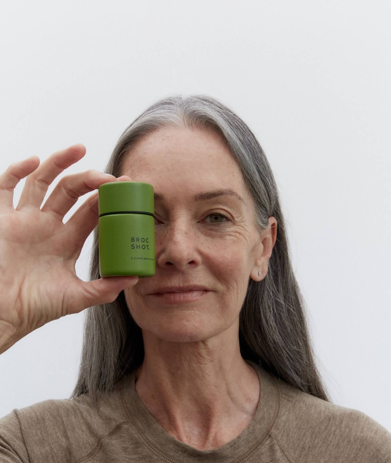 Older woman holding green container reading 'BROC SHOT. A SUPER ANTIOXIDANT' beside her left eye