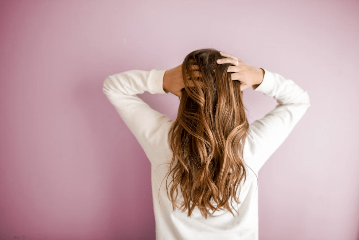 Rear view of a woman with long wavy brown hair, hands running through her hair against a pink wall.