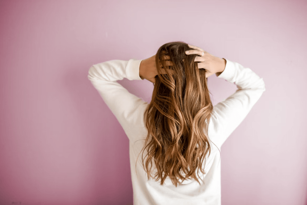 Rear view of a woman with long wavy brown hair, hands running through her hair against a pink wall.