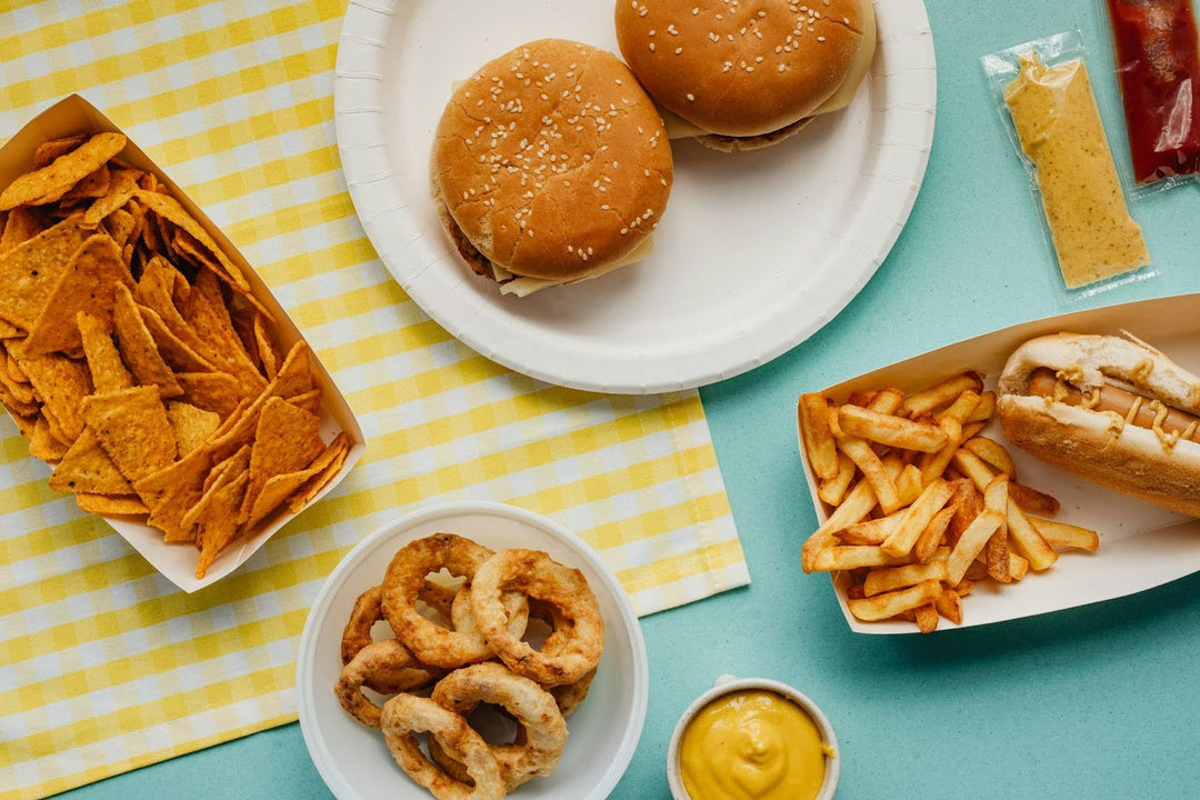 Overhead view of fast-food spread: two cheeseburgers, fries, hot dog, nacho chips, onion rings and mustard