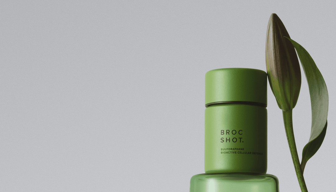 Three-quarter view of green Broc Shot bottle with matte cap and glass base beside a closed flower bud on gray background