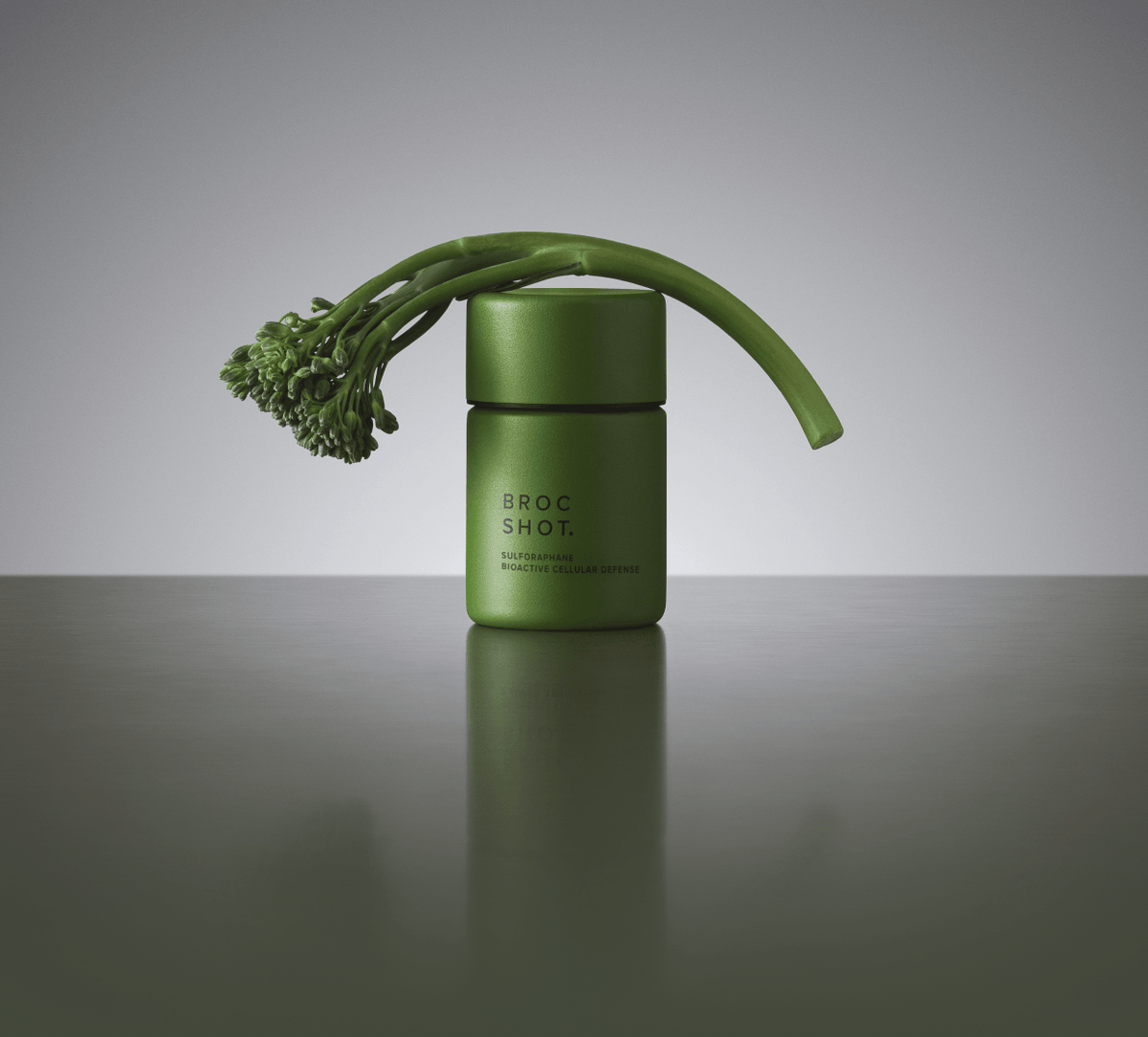 Front view of Broc Shot supplement bottle showing a matte green jar with BROC SHOT label and a curved broccoli floret draped over it, reflected on a gray surface