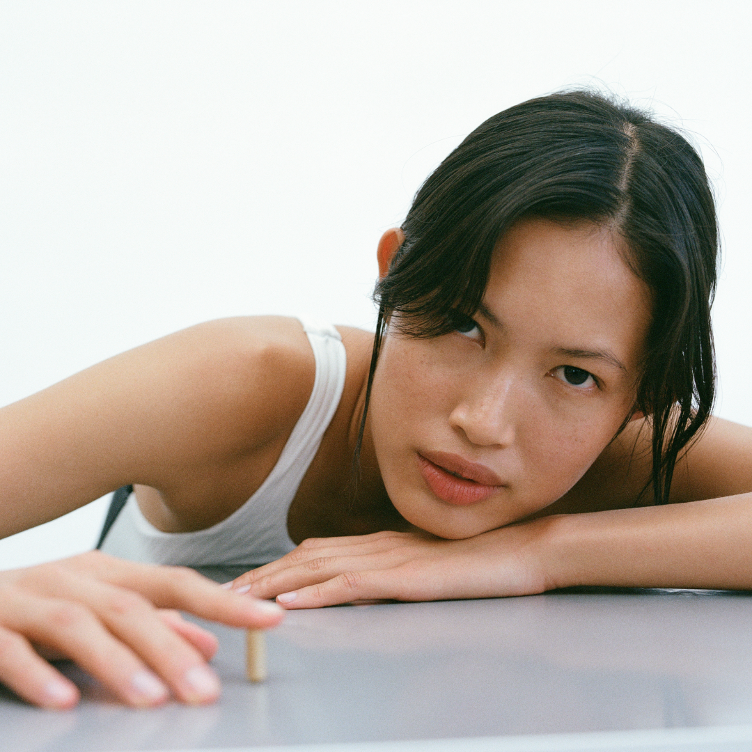 Young woman resting her head on folded arms, reaching toward a small brass screw on a table.