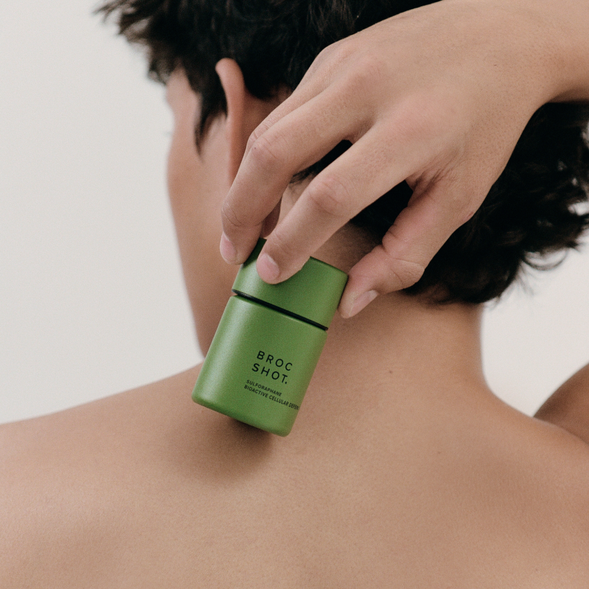 Hand holding green bottle labeled 'BROC SHOT.' and 'SULFORAPHANE BIOACTIVE CELLULAR DEFENSE' against a bare back