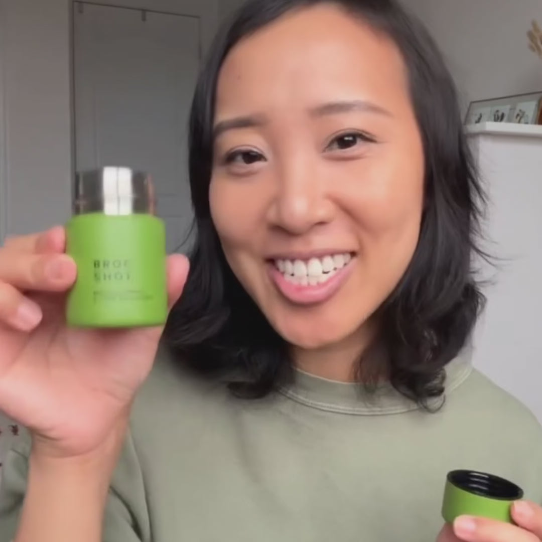 Person smiling and holding a green Broc Shot bottle up to camera against a plain light background